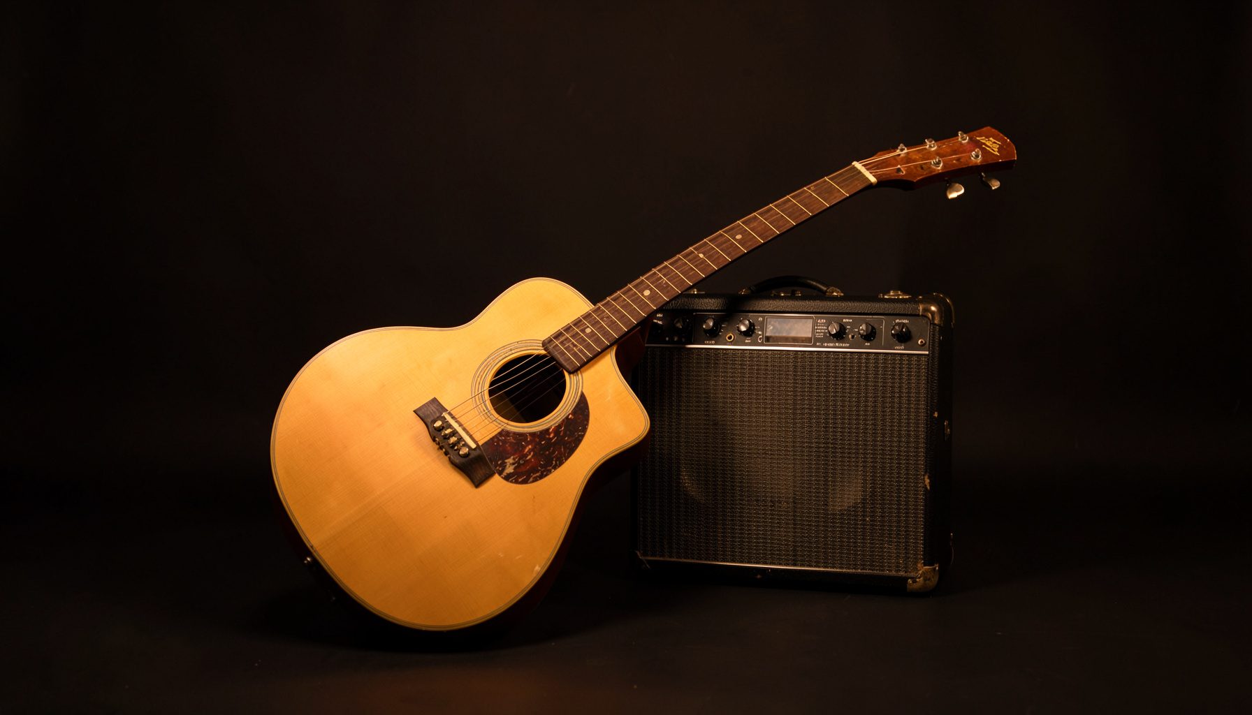 A warm, intimate studio photograph of an acoustic guitar resting against a vintage amplifier on a dark seamless backdrop, with soft amber and cream lighting creating a moody, romantic atmosphere.