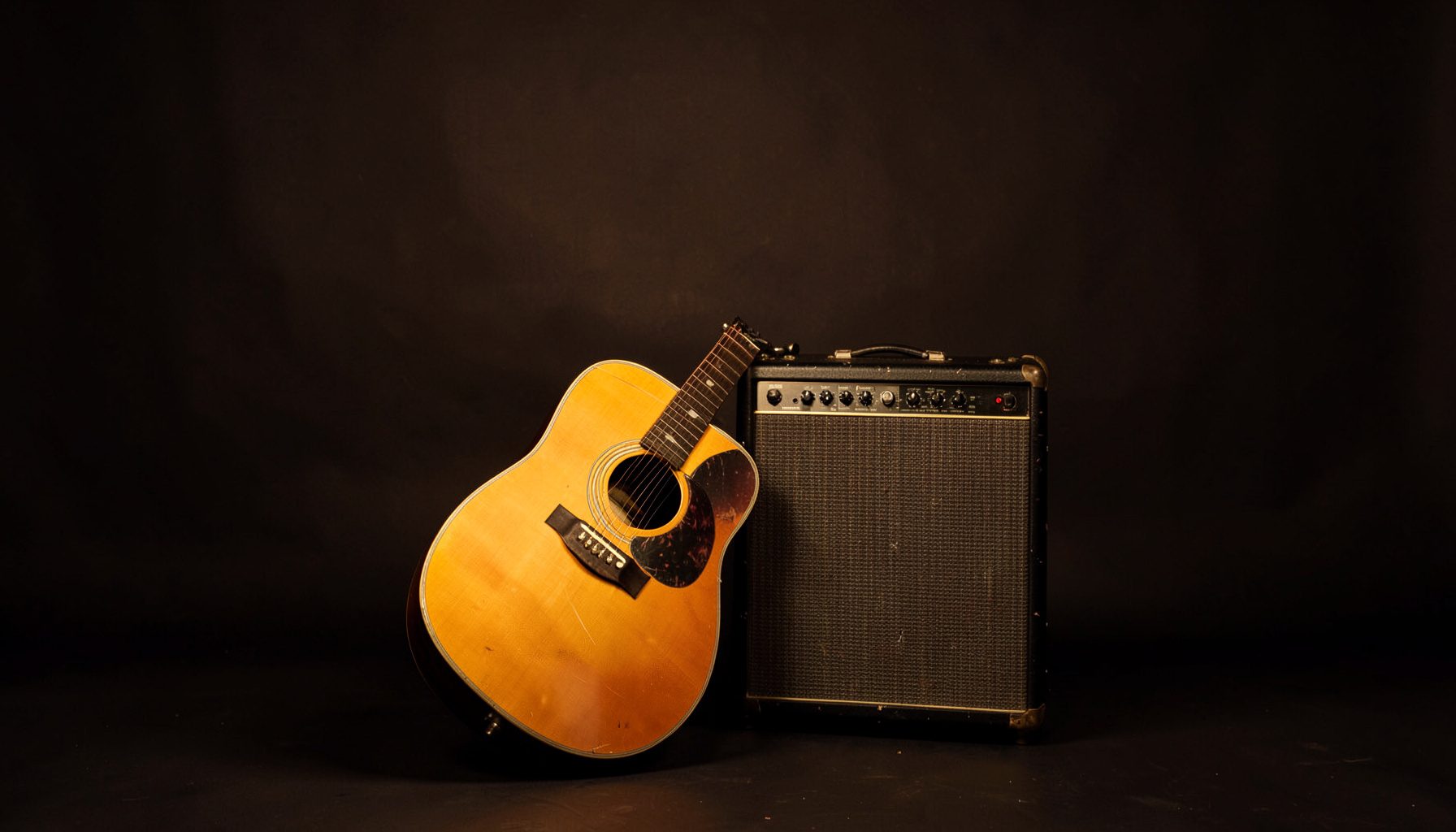 A warm, softly lit acoustic guitar resting against a weathered amplifier on a dark stage, deep browns and muted gold tones