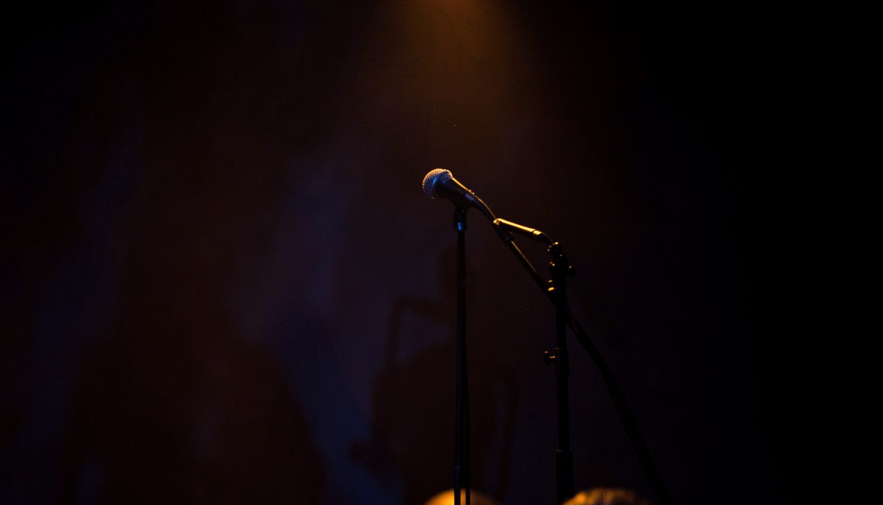A warm, intimate stage performance with soft amber and blue stage lighting, a microphone stand in the foreground, and a dark seamless backdrop.